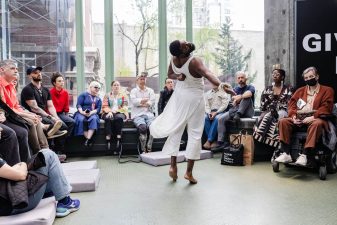 Alameda Post - Jerron Herman twirls in mid-turn dressed in a long, flowy white tunic in front of the audience in a light-filled atrium.