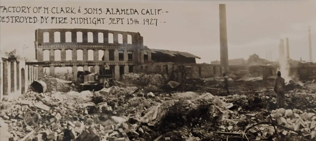 Alameda Post - a black and white photo of the smoldering remains of a factory. A person stands and observes the wreckage. In neat, old handwriting on the photo, tet reads "Factory of N. Clark & Sons Alameda Calif. Destroyed by fire. Midnight Sept 15th 1927