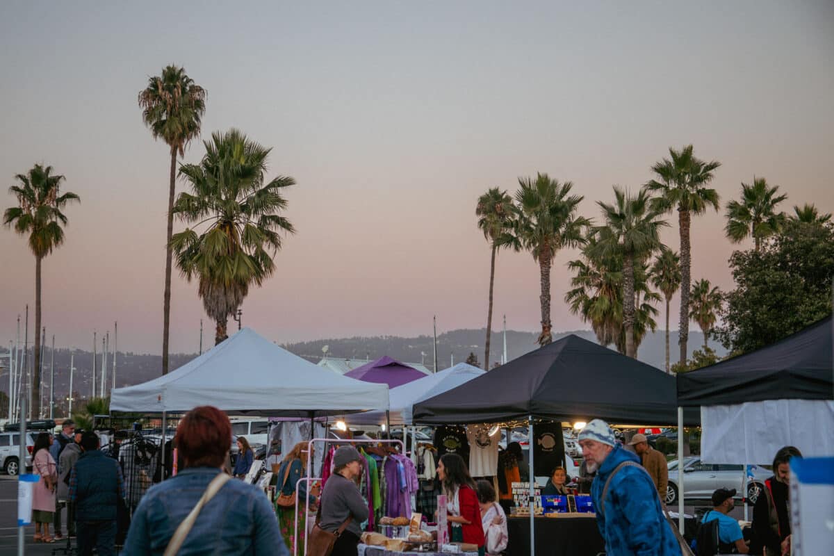 Alameda Post - Attendees shop from vendors at a market. In the background, The Oakland Hills are lit by the setting sun. 