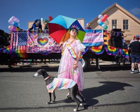 Alameda Post - a person in a pink dress with a rainbow umbrella and a dog stand in front of a pride float