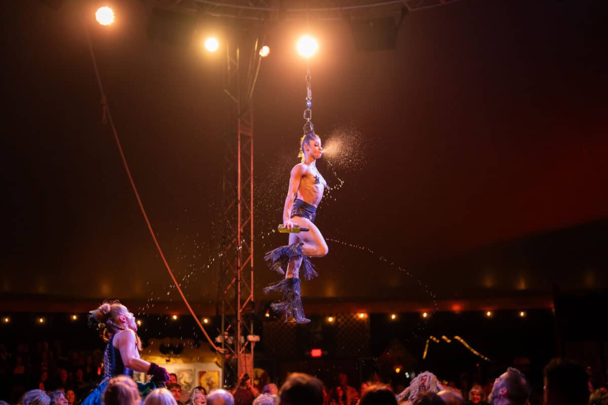 Alameda Post - an acrobat hangs by her hair while spraying water from her mouth and twirling
