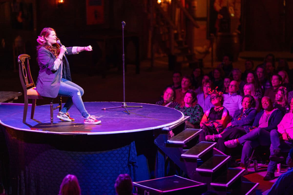 Alameda Post - A woman holds a microphone onstage at Spooky Tales. She pantomimes driving a car.