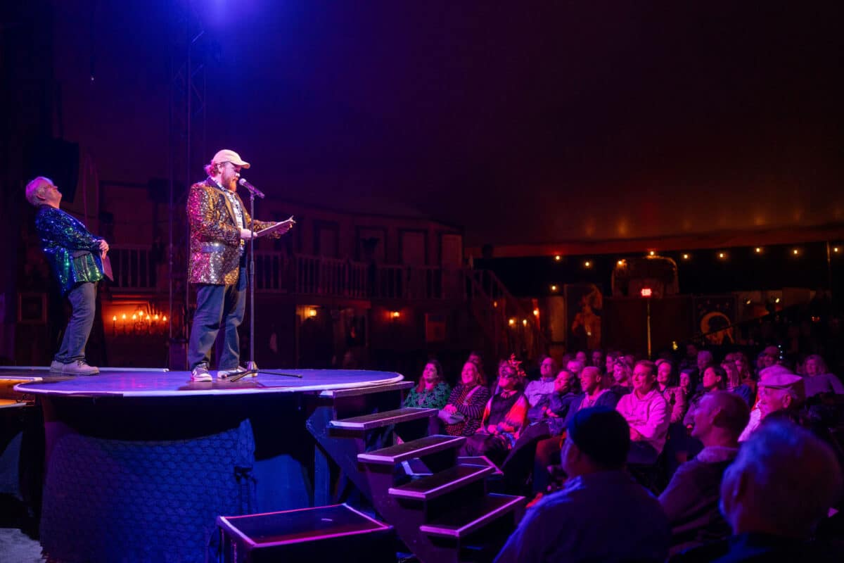 Alameda Post - Two men in sparkly vests stand on a raised platform in front of an audience. One reads off of a clipboard and speaks into a microphone.