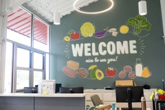 Alameda Post - A reception desk with a large colorful mural of food behind it that says "Welcome. Nice to see you!" The desk is up against an outside wall, and there is a large window that can be used to pass food from the desk to outside.