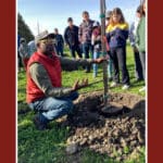 Alameda Post - a man crouches on the ground next to a sapling tree and talks to a group of people