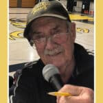 Alameda Post - Richard Bullock Sr. holds a microphone and a pencil while wearing an Alameda Hornets baseball hat in front of a basketball court