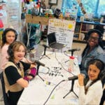 Alameda Post - Love Elementary Fourth and Fifth graders sit at a table in their classroom with podcast equipment. Three students sit with an adult woman, and they all smile at the camera.