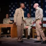 Alameda Post - two men talk heatedly onstage. A large American flag is the background