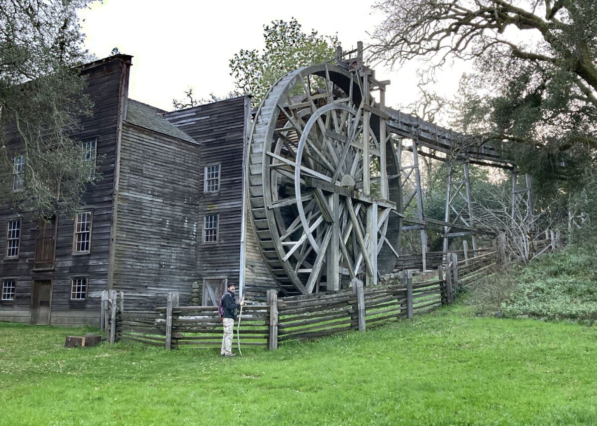 Alameda Post - A large old wooden mill with a waterwheel, and a man standing in front of it.