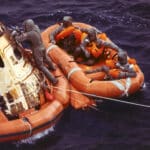 Alameda Post - three astronauts on a raft float next to a space capsule. One other person climbs on the capsule.