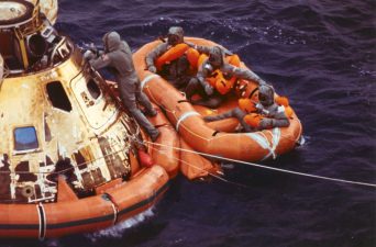Alameda Post - three astronauts on a raft float next to a space capsule. One other person climbs on the capsule.