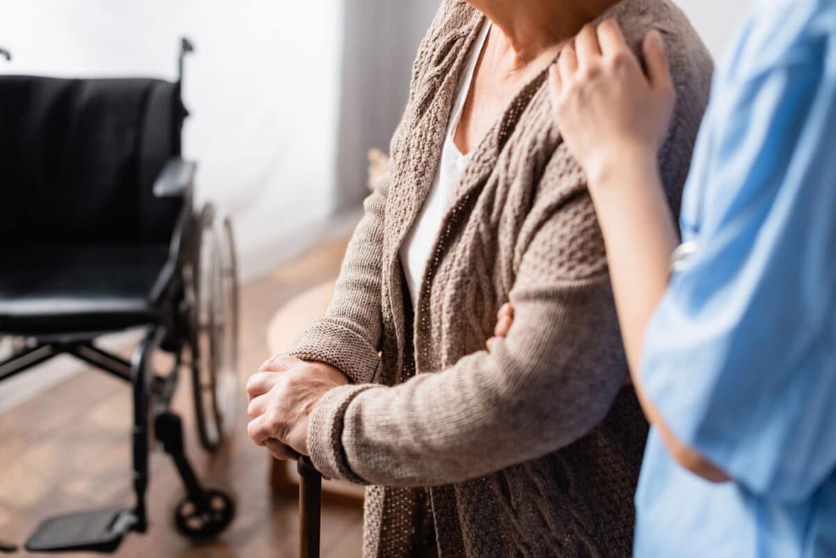 Alameda Post - a stock image of a nursing home where a resident is standing next to a wheelchair with a cane, being assisted by a healthcare worker