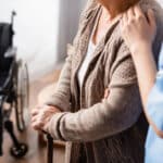 Alameda Post - a stock image of a nursing home where a resident is standing next to a wheelchair with a cane, being assisted by a healthcare worker