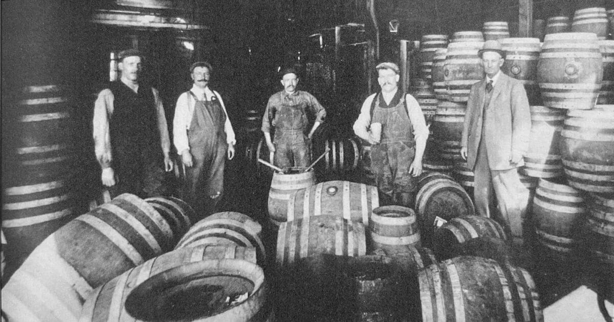 Alameda Post - Four men in work clothes and one man in a suit to the right stand amongst a lot of barrels in a black and white photo. 