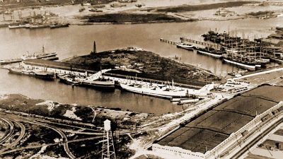 Alameda Post - A black and white photo of a piece of land jutting out into the water lined with ships. The area is called Encinal Terminals and was established and thrived thanks to the shipping industry and Calpak