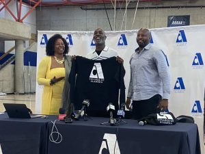 Alameda Post - Gary Payton smiles and holds up a College of Alameda sweatshirt at a press conference. He stands next to two people.