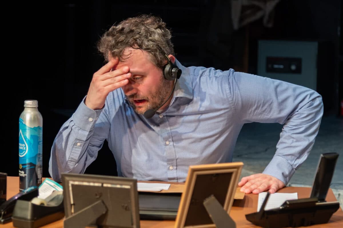 Alameda Post - The actor in Fully Committed sits at his desk wearing a call headset. His mouth is open as if speaking or sighing, and his had is in his hand. 