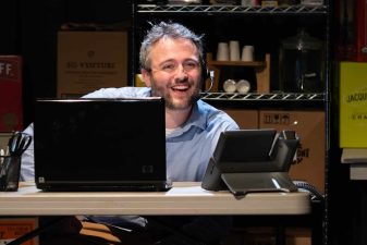 Alameda Post - An actor in Fully Committed sits behind a desk wearing a call headset and smiles.