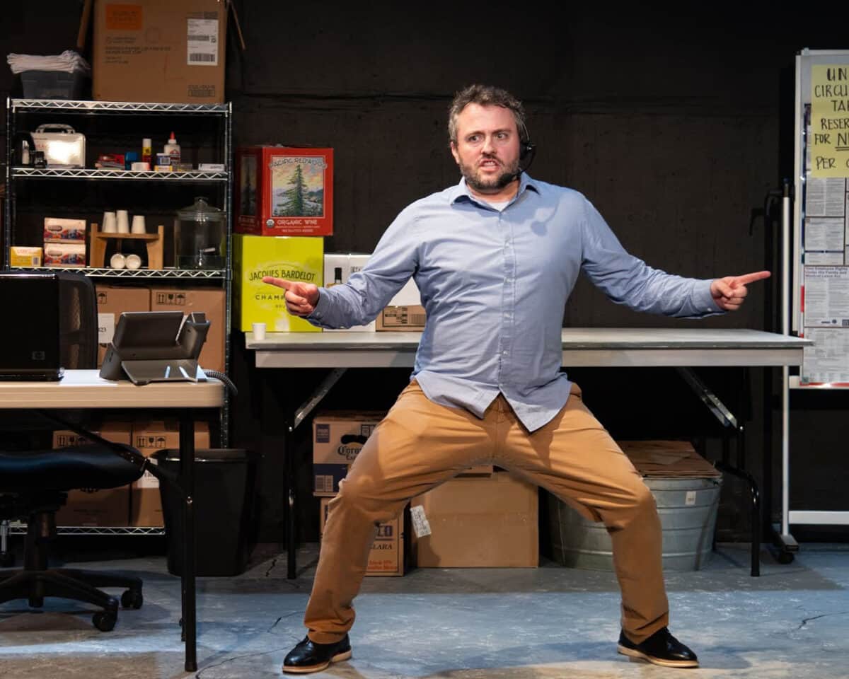 Alameda Post - An actor in khakis and a button up, wearing a call headset, takes a wide standing stance onstage and points his fingers to the left and right while grimacing. 