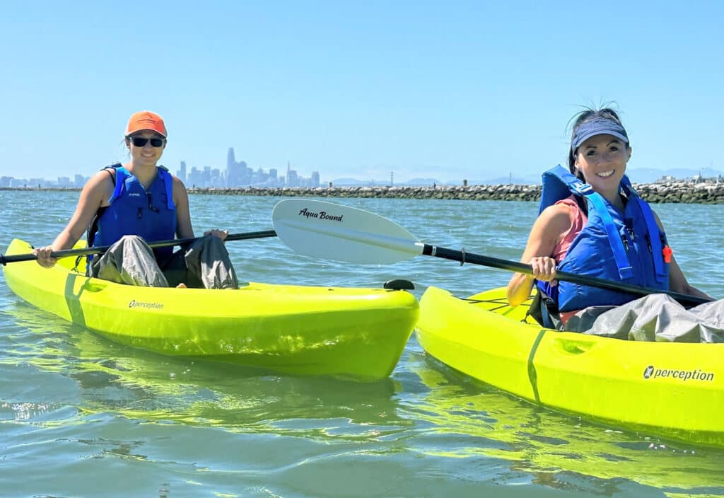 Alameda Post - two women in kayaks float in Ballena Bay with San Francisco in the distance on a Sea Trek tour