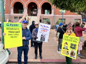 Alameda Post - protesters on President's Day at Alameda City Hall hold signs that say things like "Greedy heartless Trump Musk" and "Stop Cutting Firefighter Jobs"