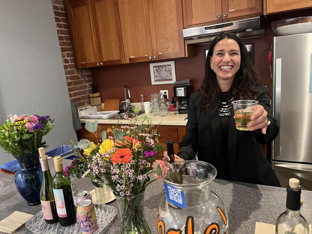 Alameda Post - A smiling woman holds out a glass of wine towards the camera.