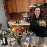 Alameda Post - A smiling woman holds out a glass of wine towards the camera.