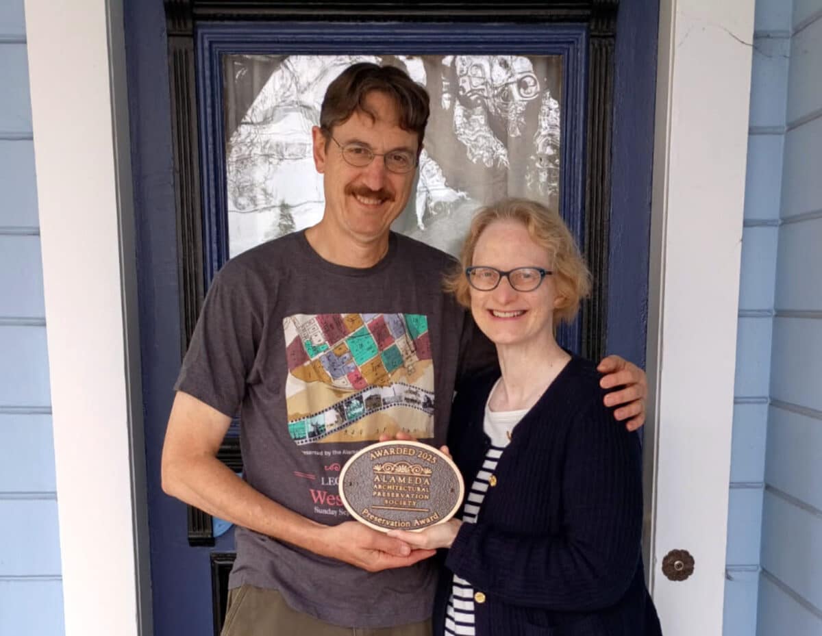Alameda Post - a man and woman stand together in front of a front door and smile. They hold a plaque from the AAPS.