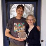 Alameda Post - a man and woman stand together in front of a front door and smile. They hold a plaque from the AAPS.