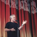 Alameda Post - a man talks into a microphone onstage in front of a large red curtain