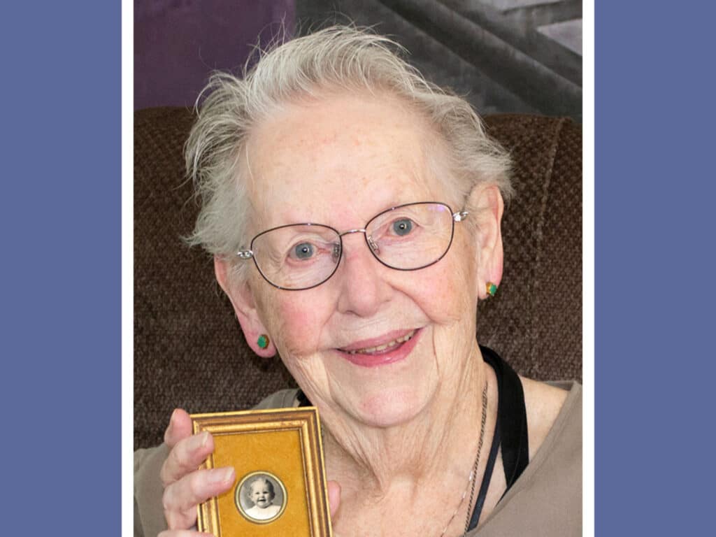 Alameda Post - an older woman, Joan Mary Spiersch Brammer, smiles and holds a black and white portrait of a young girl