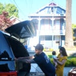 Alameda Post - The May family packs a box into the trunk of a vehicle. One child is in their mother's arms, and another runs around the yard.