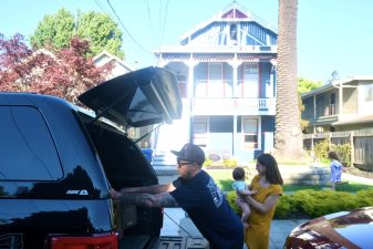 Alameda Post - The May family packs a box into the trunk of a vehicle. One child is in their mother's arms, and another runs around the yard.