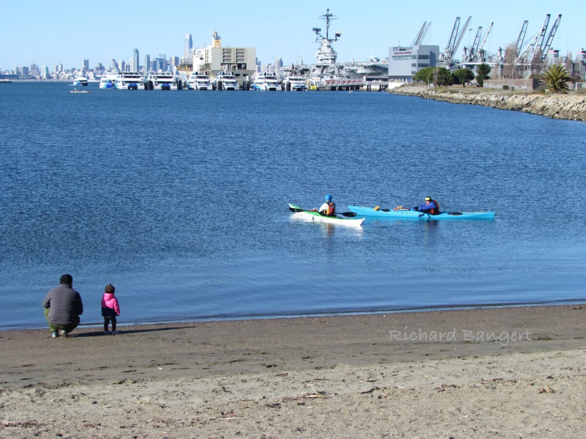Alameda Post - A parent and young child sit on Encinal Beach and watch kayakers. 
