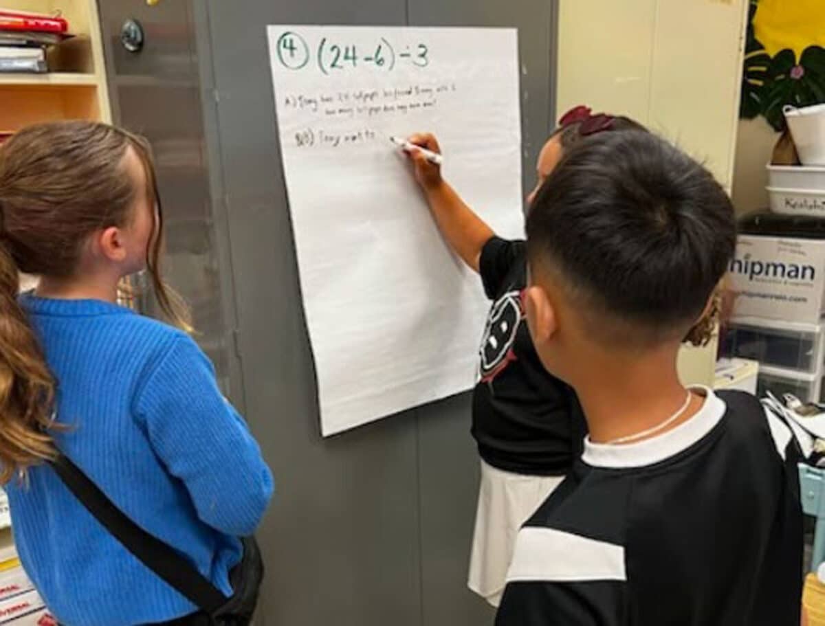 Alameda Post - Three fourth graders stand around a large piece of paper attached to the wall and work on a math problem together. 