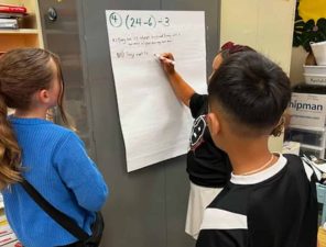 Alameda Post - Three fourth graders stand around a large piece of paper attached to the wall and work on a math problem together.