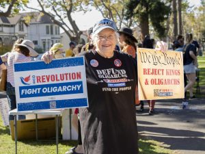 Alameda Post - A woman stands in Chochenyo park holding protest signs that say "We the People Reject Kings & Oligarchs" and "Our Revolution. Fight Oligarchy."