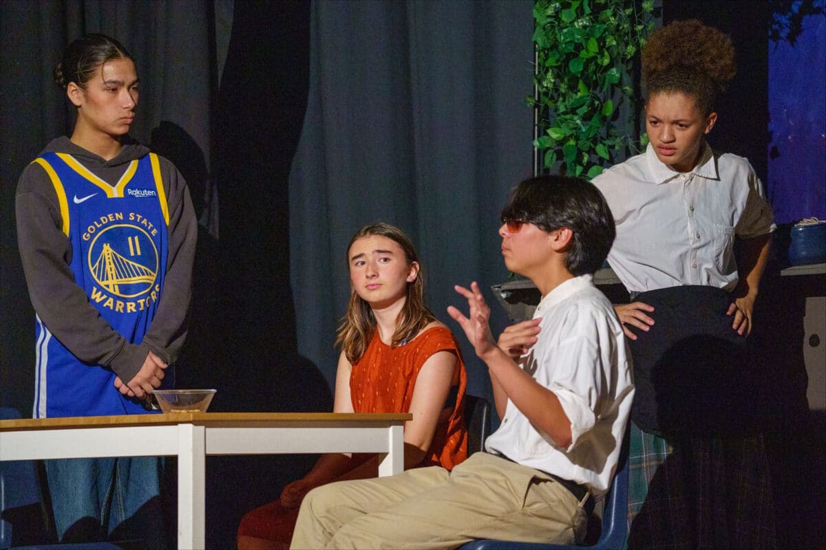 Alameda Post - Four actors sit and stand around a table. 