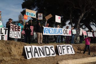 Alameda Post - A line of beach protestors hold signs that say things like "Hands Off!" " Venezuela's Oil!" and "Free Epstein."