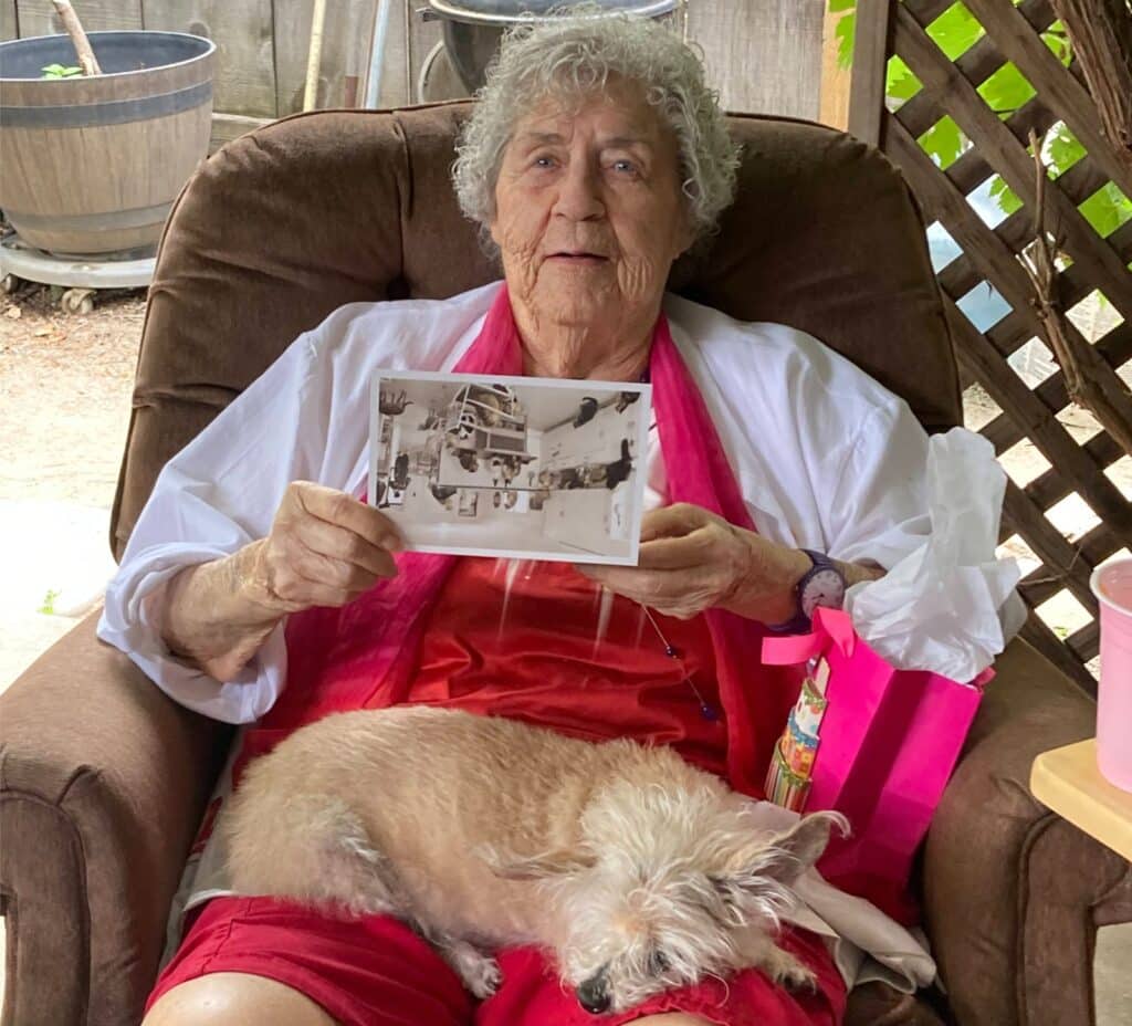 Alameda Post - Juanita Gex sits in a chair with a dog on her lap and holds a photo