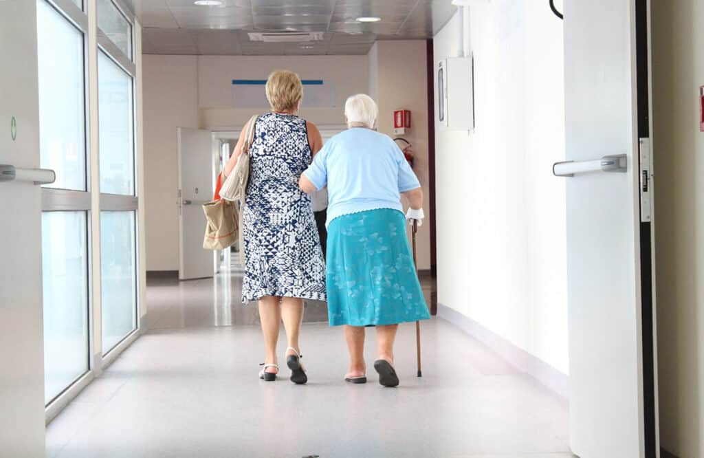 Alameda Post - two women walk together down a doctor's office or hospital hallway. One uses the other's arm for support
