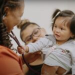 Alameda Post - a young child smiles at a woman doctor and grabs the stethescope