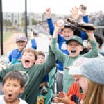 Alameda Post - in the stands of an Oakland Ballers game, kids cheer and celebrate while one holds a baseball