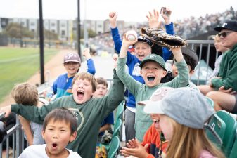 Alameda Post - in the stands of an Oakland Ballers game, kids cheer and celebrate while one holds a baseball