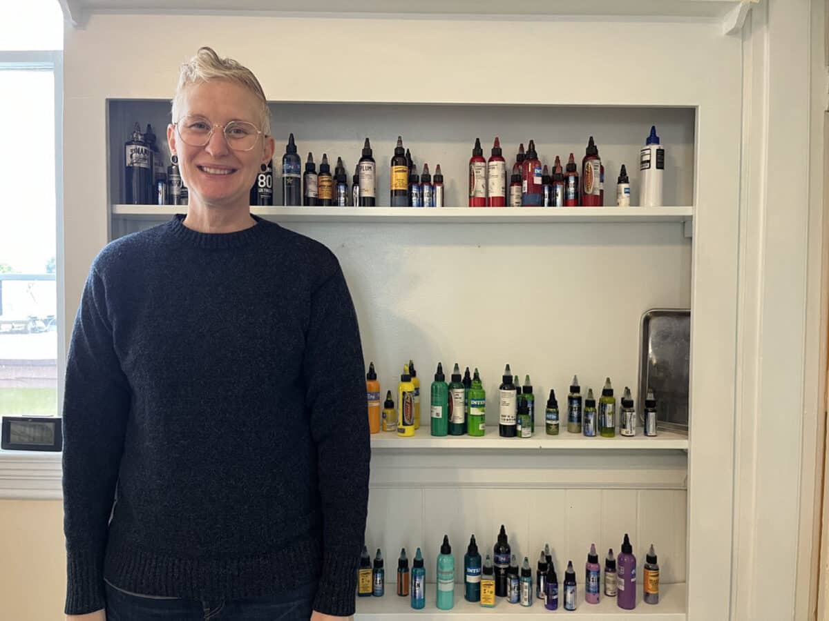 Alameda Post - A person in a blue sweater with short blond hair smiles at the camera in front of a wall of tattoo ink colors. 
