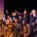 Alameda Post - the cast of Urinetown sings together in a formation on stage. A row of actors in clean suits stands behind a row of actors wearing disheveled clothing. They all wave small pieces of paper.