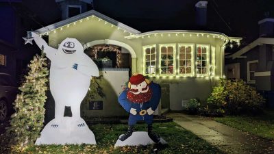 Alameda Post - large paintings of Yukon Cornelius and Bumble the Abominable Snowmonster of the North stand in the front yard of a house. The artwork is lit, and the house is covered in lights too.